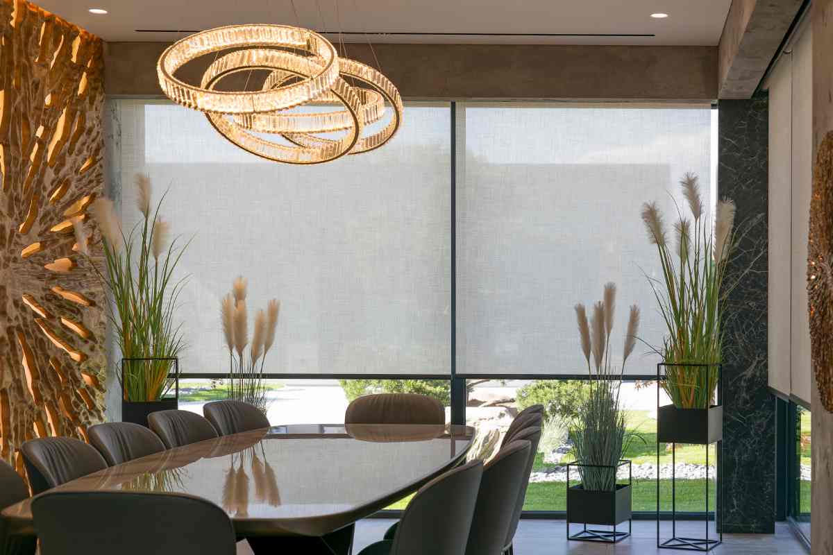 Solar shades blocking light in an upscale dining room with a chandelier and potted plants at Blinds Plus Designs near Huntington Beach, CA